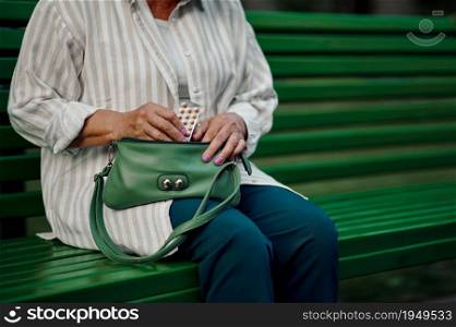 Granny takes the pills out of her bag on the bench in summer park. Aged people lifestyle. Pretty grandmother having fun outdoors, old female person on nature. Granny takes pills out of her bag in summer park