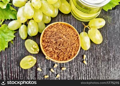 Grape seed flour in a bowl, oil in a jar and green grapes on dark wooden board background from above