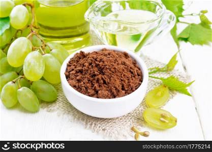 Grape seed flour in a bowl on a burlap napkin, oil in a jar and gravy boat, green grapes on the background of light wooden board