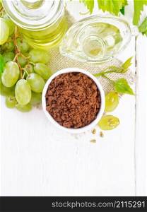 Grape seed flour in a bowl on sacking, oil in a jar and gravy boat, green grapes on the background of light wooden board from above