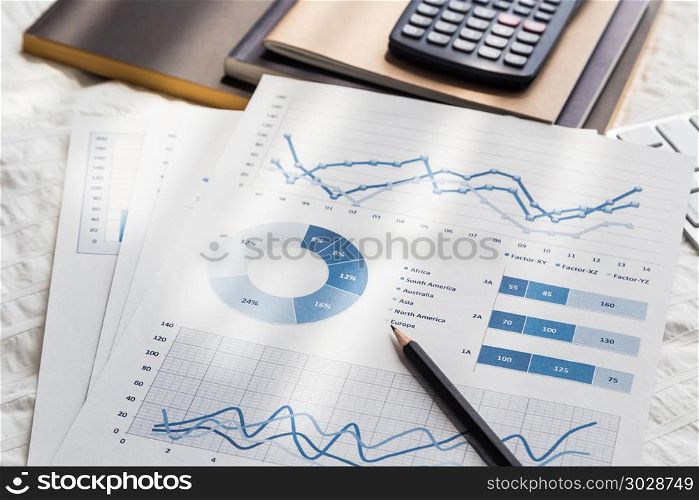 Graphs analysis documents work at home on bed.. Blue chart graph data analysis documents, pencil, calculator, notebooks on wrinkle white duvet bedding fabric. Morning daylight in bedroom. Freelance job work at home, business, education concepts.