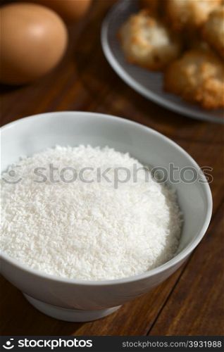 Grated coconut in bowl with coconut macaroons and eggs in the back, photographed with natural light (Selective Focus, Focus one third into the grated coconut)