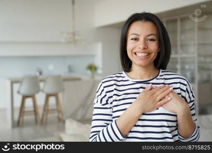 Grateful happy young woman holding hands on chest feeling thankful≠ss, love, standing at home. Touched thankful fema≤homeow≠r housewife expressing gratitude, appreciation, thanking, smiling.. Touched thankful woman thanks smiling, holding hands on chest feeling love, appreciation at home