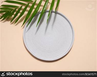 Gray empty decorative plate and green palm leaf on a beige background, product and cosmetic display stage, top view