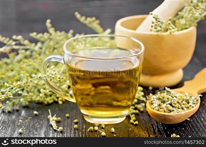 Gray wormwood herbal tea in a glass cup, dry sagebrush flowers in spoon, fresh flowers in a mortar and on table against black wooden board