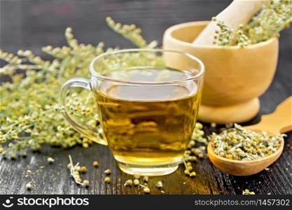 Gray wormwood herbal tea in a glass cup, dry sagebrush flowers in spoon, fresh flowers in a mortar and on table against black wooden board