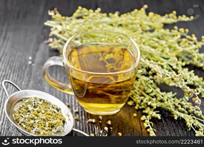 Gray wormwood herbal tea in a glass cup, fresh flowers and metal strainer with dried flowers sagebrush on wooden board background