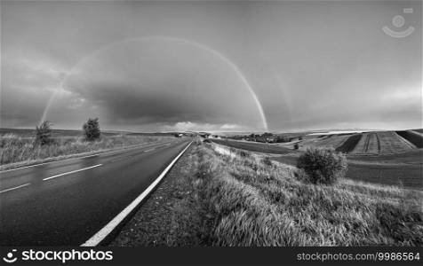 Grayscale. Spring rapeseed and small farmlands fields after rain evening view, cloudy pre sunset sky with rainbow and rural hills. Seasonal, weather, climate, eco, farming, countryside beauty concept.