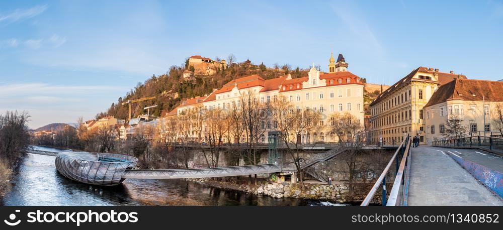 Graz, Styria Austria - 20.01.2019: Panorama view at Mur river, Murinsel on bridge, Schlossberg hill with fortress and clock-tower Uhrturm. Travel destination.. Panorama view at Mur river, Murinsel on bridge, Schlossberg hill with fortress and clock-tower