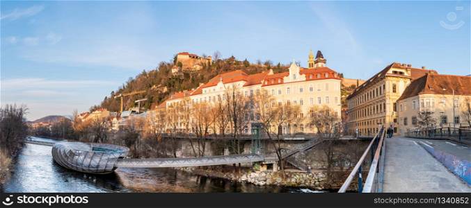 Graz, Styria Austria - 20.01.2019: Panorama view at Mur river, Murinsel on bridge, Schlossberg hill with fortress and clock-tower Uhrturm. Travel destination.. Panorama view at Mur river, Murinsel on bridge, Schlossberg hill with fortress and clock-tower