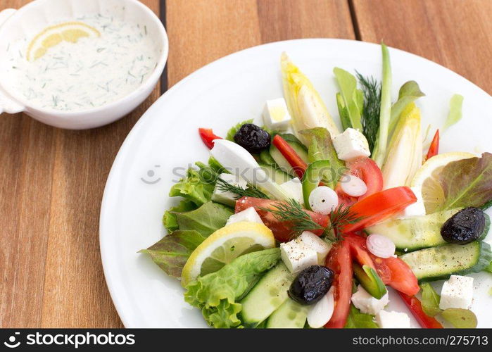 Greek salad with feta, tomatoes, cucumber, peppers and black olives. With bowl of sauce on a wooden table.