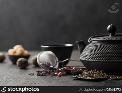 Green and black loose tea with sugar and Chinese teapot with spoons and cup with infuser and spoons on black background