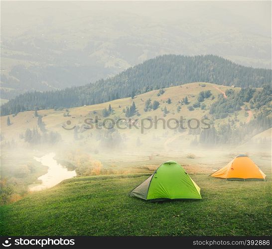 Green and orange tents on a plain in the mountains on a foggy morning. Summer landscape. The concept of freedom of solitude and travel.. Green and orange tents on plain in mountains on foggy morning