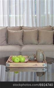 Green apple and ceramic cup in wooden tary on glass top table next to gray color sofa set