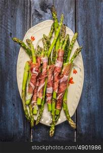 Green asparagus wrapped in ham in plate on rustic blue wooden background, top view