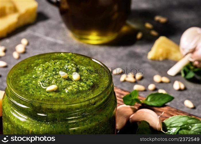 Green basil pesto sauce in a glass jar, decorated with pine nuts. Close-up, selective focus on italian sauce, ingredients on gray background