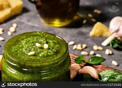 Green basil pesto sauce in a glass jar, decorated with pine nuts. Close-up, selective focus on italian sauce, ingredients on gray background