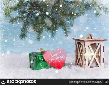 Green box and a wooden lantern in the snow under the falling snow. With fir branches on the background and a congratulatory text