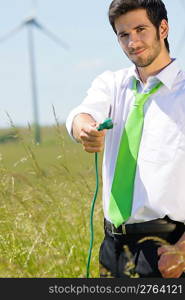 Green energy - young businessman hold plug in field with windmill