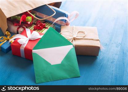 Green envelope with blank white paper leaning against a bunch of colorful presents, that come out of a brown shopping bag, on a blue wooden background.