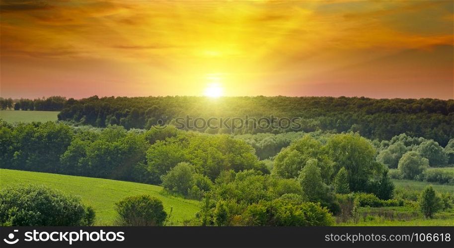 Green fields and a bright sunrise over the horizon. The hills are covered with trees and shrubs. Wide photo. Agricultural landscape.