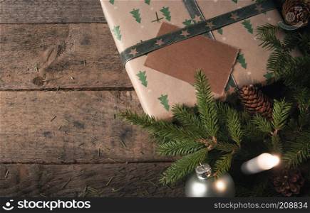 Green fir twigs, pine cones, and Christmas balls, near a gift wrapped in classic brown paper with trees, and a lit candle, on a vintage wooden table.