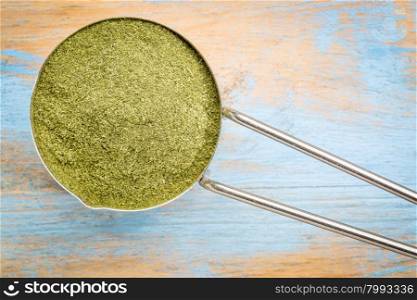 green freeze-dried organic wheat grass powder, nutritional supplement on a metal measuring scoop against painted grunge wood - top view