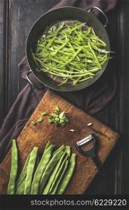 Green french beans cooking. Slices Green french beans in cooking pot and vegetable peeler on dark rustic wooden background, top view