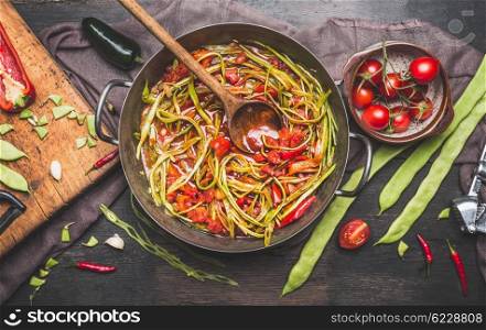 Green French beans meal preparation with wooden spoon. Green French beans in cooking pot with tomatoes sauce and ingredients on dark rustic background, top view. Vegetarian food concept