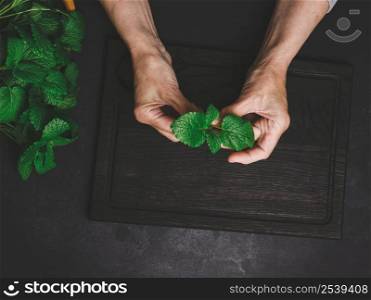 Green fresh mint leaves on a wooden board and two female hands on a black wooden table, top view