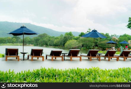 Green grass on blur wooden beach chair and beach umbrella beside swimming pool at luxury hotel near the mountain. Summer vacation travel. Tropical holiday. Eco-friendly and sustainable hotel or resort