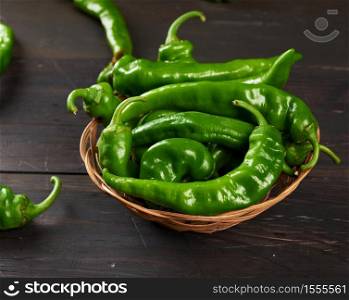 green hot pepper pods in a round wicker basket on a brown wooden table, top view