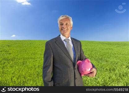 Green investment concept shot of a man or businessman in suit holding a pink piggy bank in a green field with a bright blue sky.