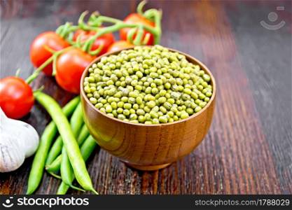 Green lentils mung in a bowl, pods of beans, garlic and red tomatoes on the background of a dark wooden board
