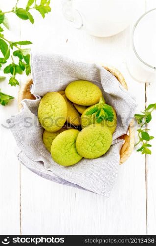 Green mint cookies on a napkin in wicker basket, milk in a glass and a jug on the background of light wooden board from above
