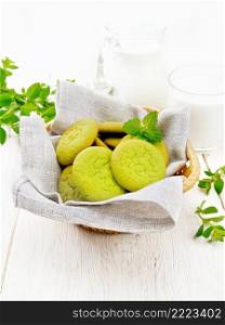 Green mint cookies on towel in wicker basket, milk in a glass and a jug on wooden board background