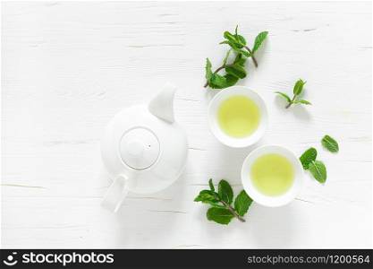 Green mint tea with fresh leaves in cups and teapot overhead on white wooden table, healthy warming drink, antioxidant beverage, top view