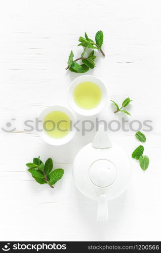 Green mint tea with fresh leaves in cups and teapot overhead on white wooden table, healthy warming drink, antioxidant beverage, top view