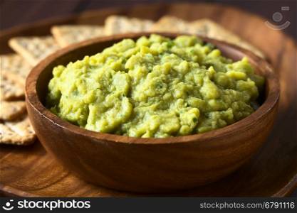 Green pea and parsley dip or spread in wooden bowl with soda crackers in the back, photographed with natural light (Selective Focus, Focus in the middle of the dip)