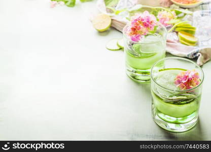 Green refreshing summer drinks with ice in glasses on light table with ingredients and flowers. Fresh  iced lemonade with cucumber and herbs. Copy space for your design