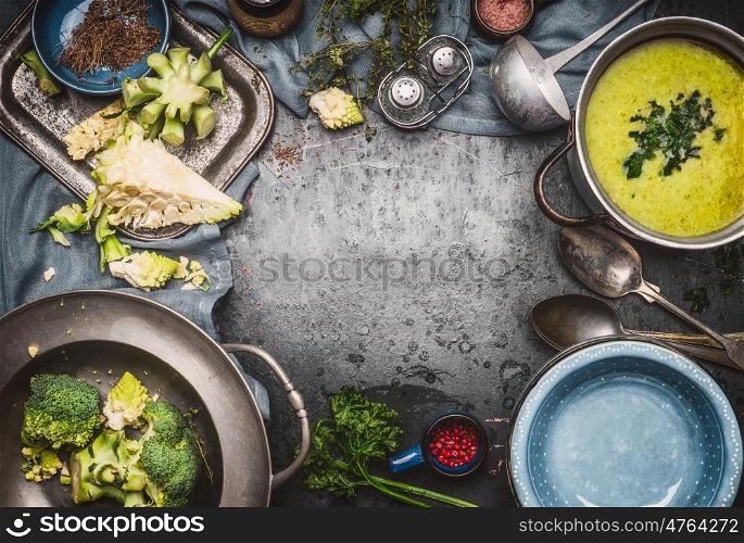 Green Romanesco and broccoli soup with cooking ingredients,kitchen tools, ladle , bowls and spoons on dark rustic background, top view, frame. Healthy and vegetarian food or diet nutrition concept.
