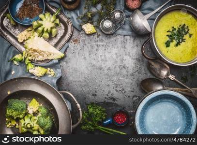 Green Romanesco and broccoli soup with cooking ingredients,kitchen tools, ladle , bowls and spoons on dark rustic background, top view, frame. Healthy and vegetarian food or diet nutrition concept.
