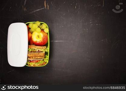 Green school lunch box with sandwich, apple, grape and carrot close up on black chalkboard background. Healthy eating habits concept. Flat lay composition (from above, top view). Space for text