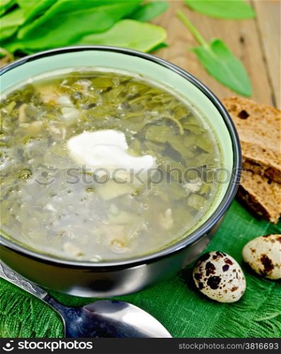 Green soup of sorrel and spinach in a bowl with sour cream on a napkin, quail eggs, bread, spoon on a wooden boards background