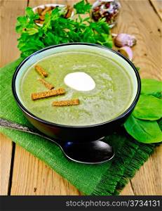 Green soup puree in a bowl with a spoon on a napkin and spinach, parsley, croutons, garlic and pepper on a wooden board