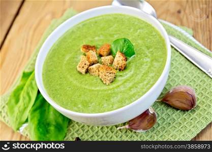 Green soup puree in a bowl with green spinach leaves, spoon, garlic on a napkin on a wooden boards background