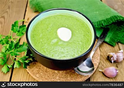 Green soup puree in bowl with a spoon on a stand, napkin, parsley, garlic and pepper on a wooden board