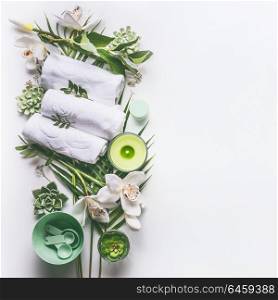 Green spa or wellness background with towels, candle, tropical leaves , orchid flowers, succulents and body and face care tools and accessories on white desktop, top view with copy space.
