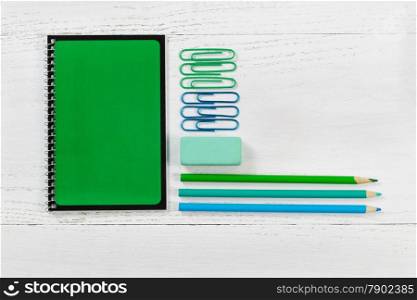 Green spiral notepad with pencils, eraser, and paper clips on desktop. Pattern in colors of green and blue with a white wooden background. Educational or business concept. &#xA;