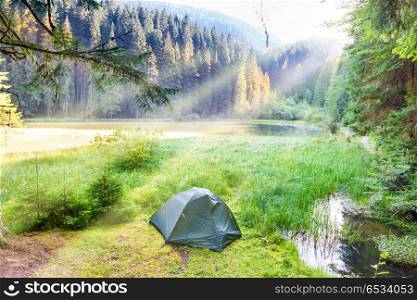 Green tent near forest lake in the mountains with blue water and morning light. Green tent near forest lake
